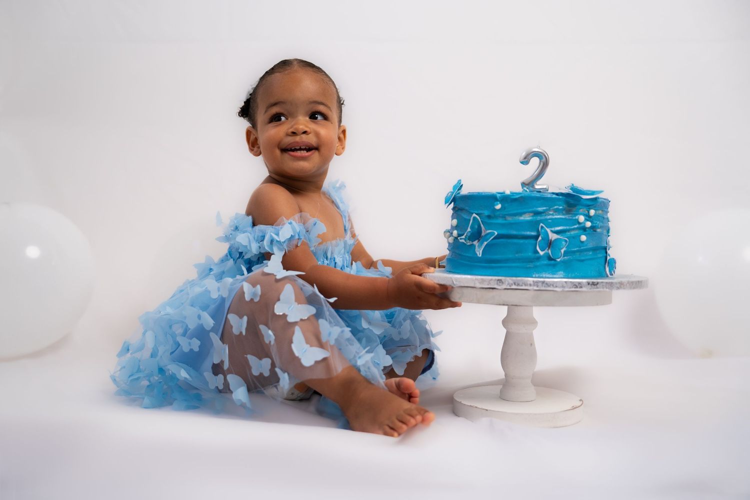 Toddler in blue tulle dress sits beside a blue birthday cake on a white pedestal.