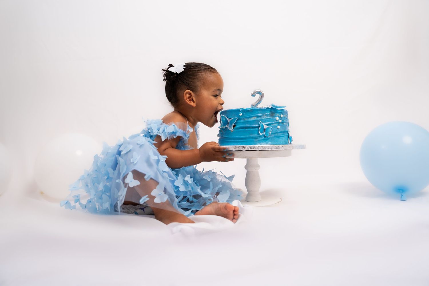 Baby in blue outfit crawling towards birthday cake and balloon on white backdrop.