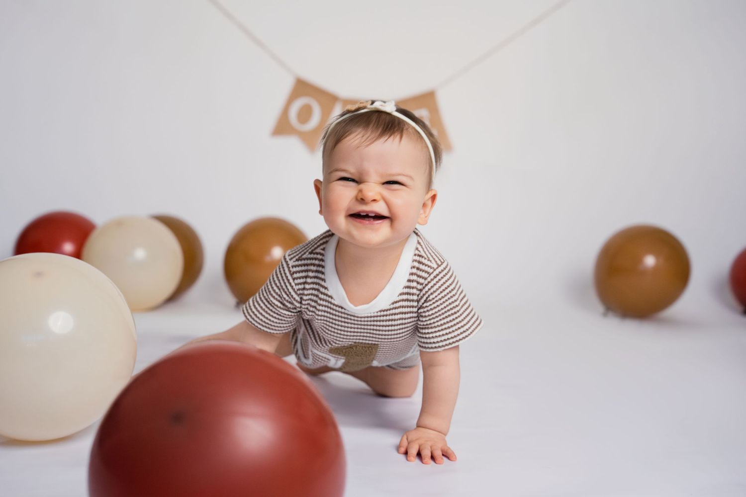 Smiling baby crawling on white floor surrounded by brown and cream balloons during a first birthday cake smash photoshoot.