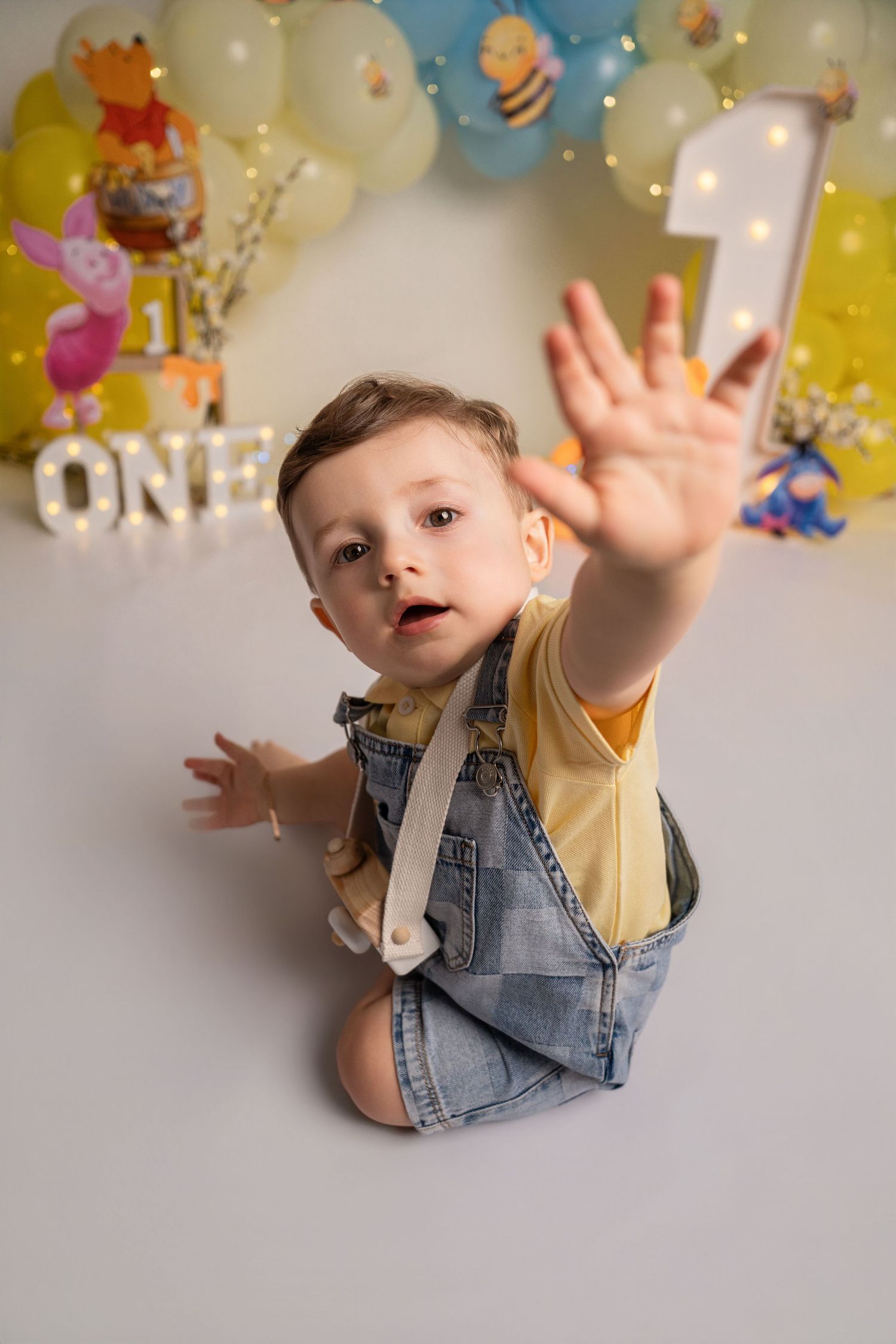 Baby boy in denim overalls reaches toward camera at a Winnie the Pooh first birthday cake smash photo session.