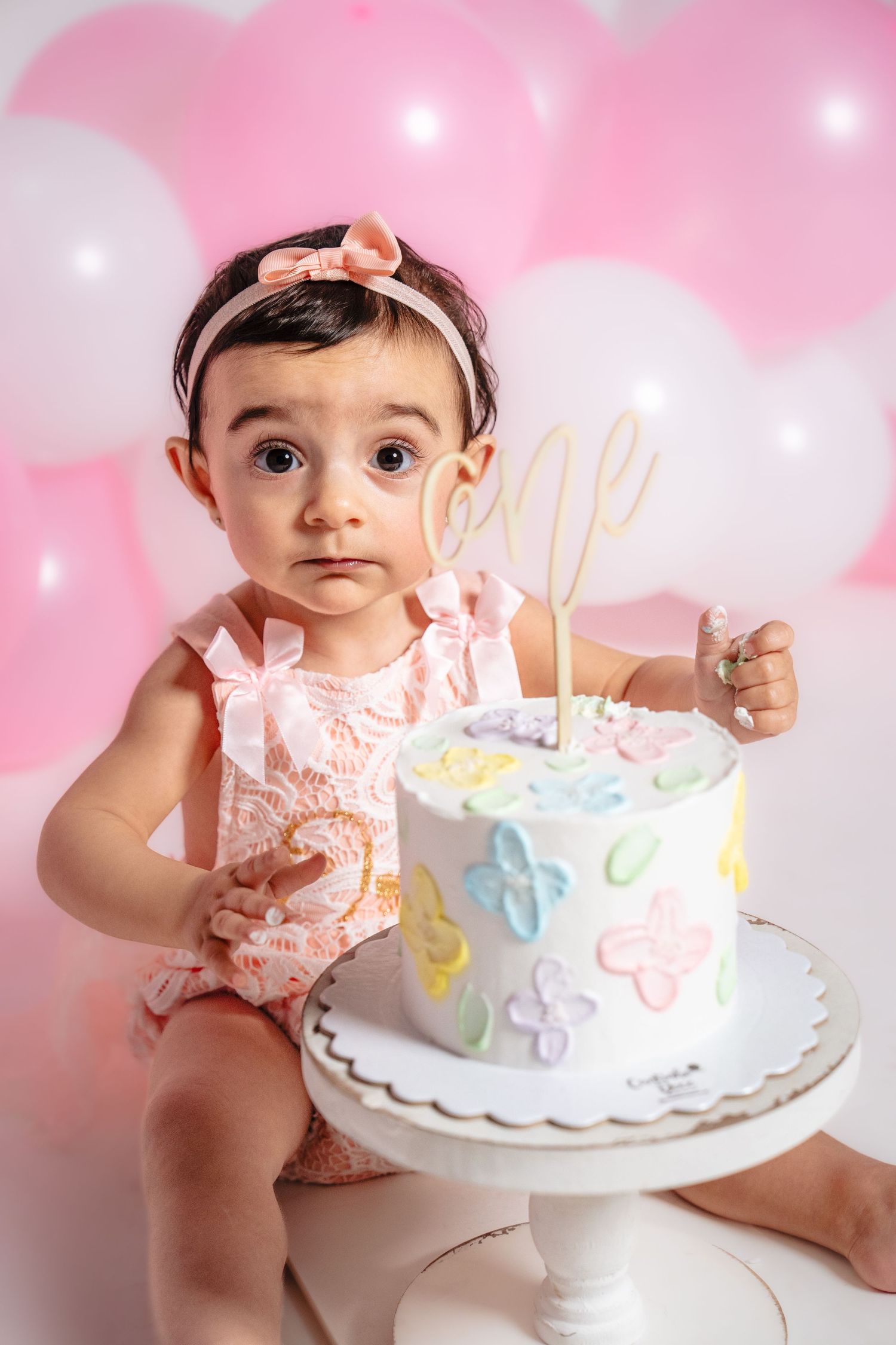 Baby girl in pink outfit sits beside white birthday cake with pastel decorations.