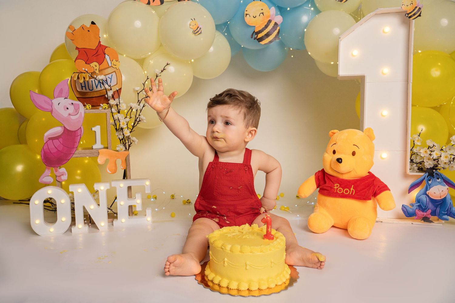 Baby boy in red romper smashing yellow cake at Winnie the Pooh first birthday party with balloon arch.