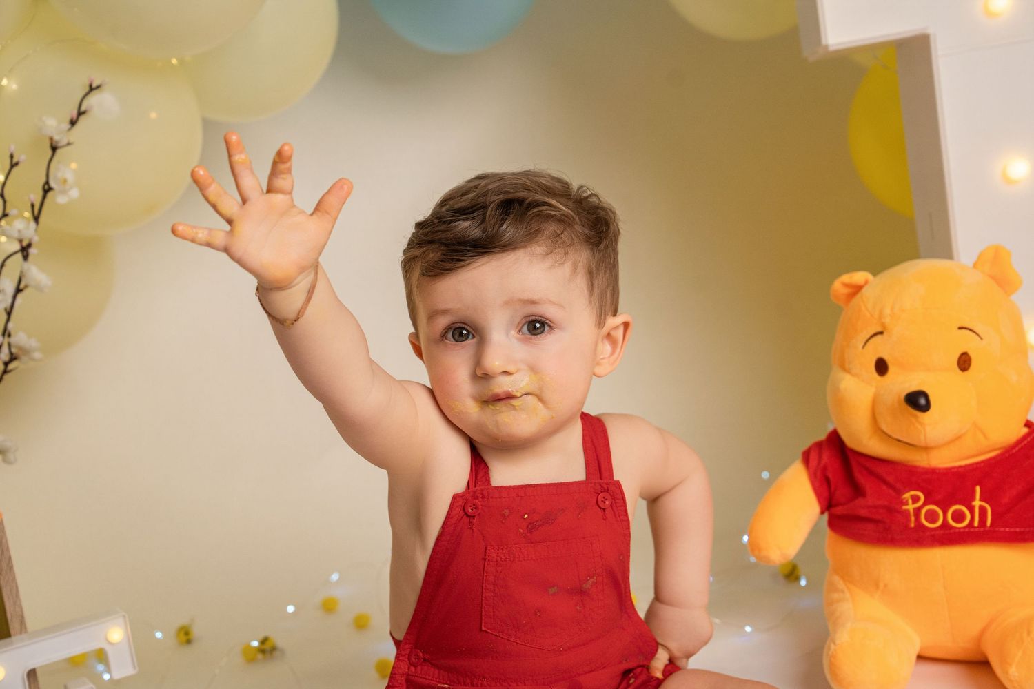 Smiling baby in red overalls waving hand next to Winnie the Pooh stuffed toy with yellow balloon decorations.