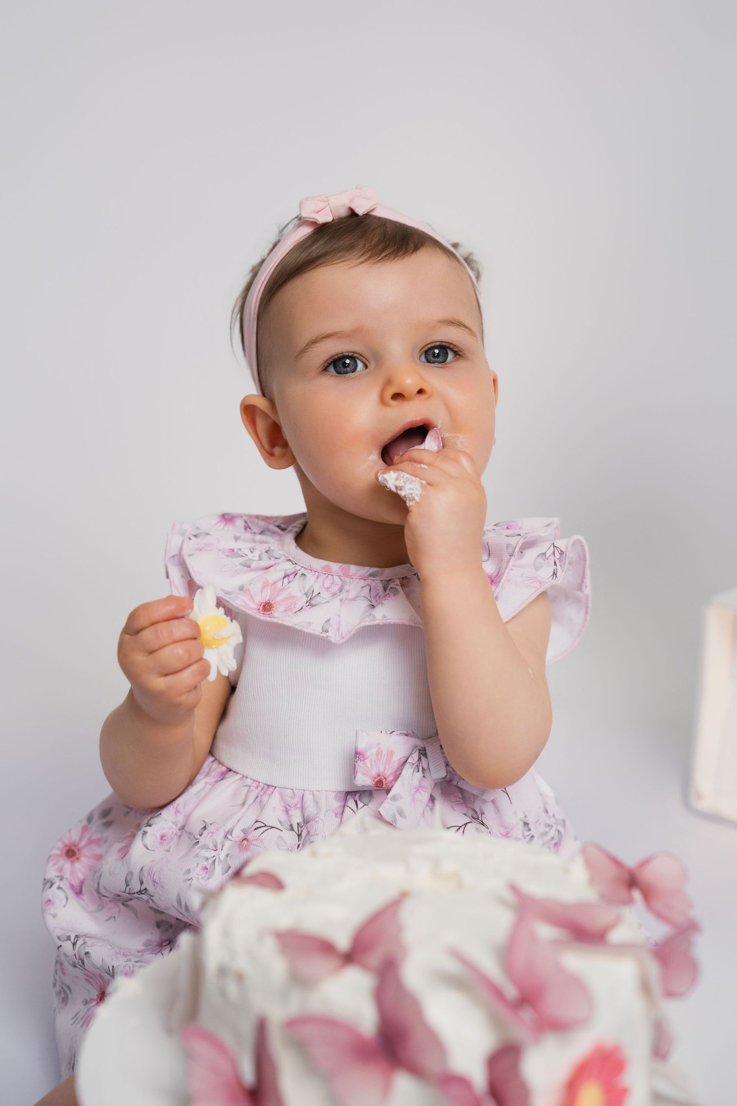 Baby girl in pink floral dress eating birthday cake with hands, celebrating first birthday with white decorated cake.