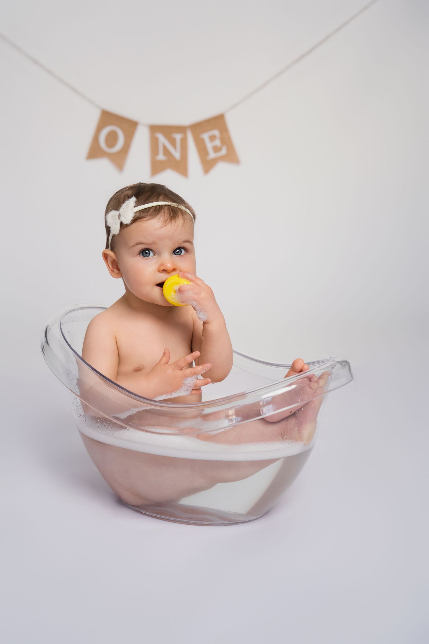 Baby girl in clear tub holding yellow duck toy, celebrating first birthday with ONE banner backdrop.