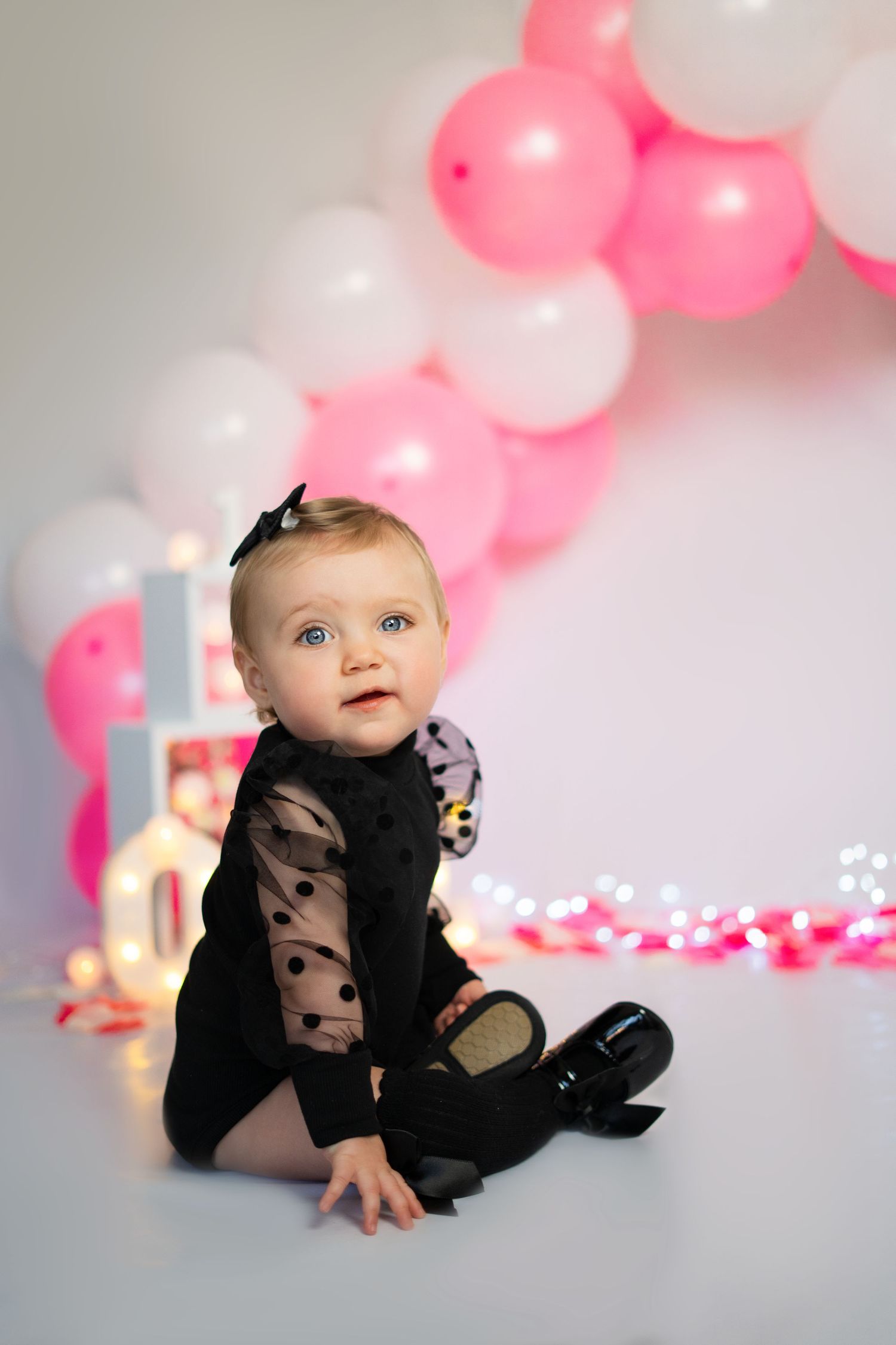 Baby in black outfit sits on floor surrounded by pink and white balloons and twinkle lights.