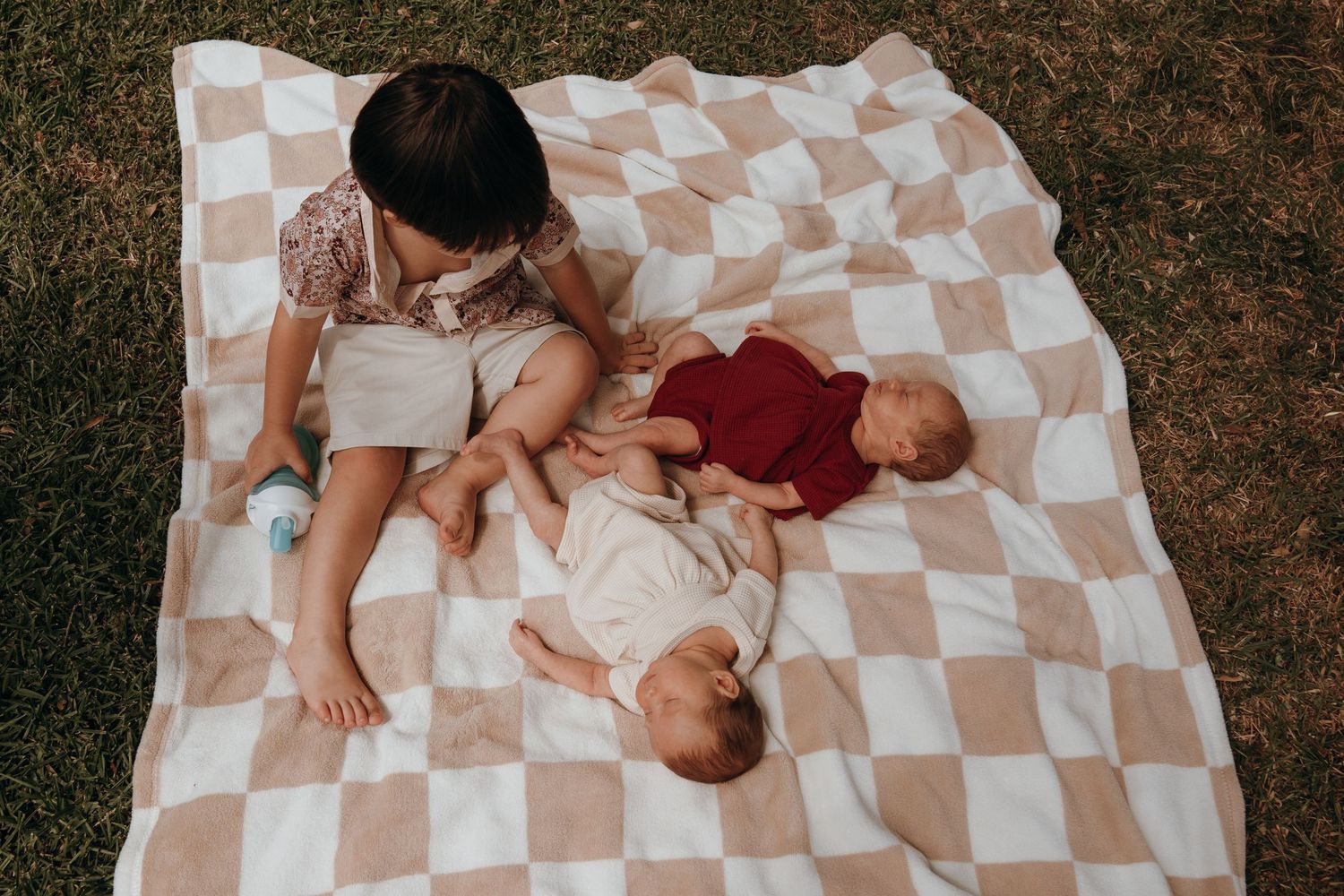Children resting together on a checkered blanket laid out on grass during outdoor leisure time.