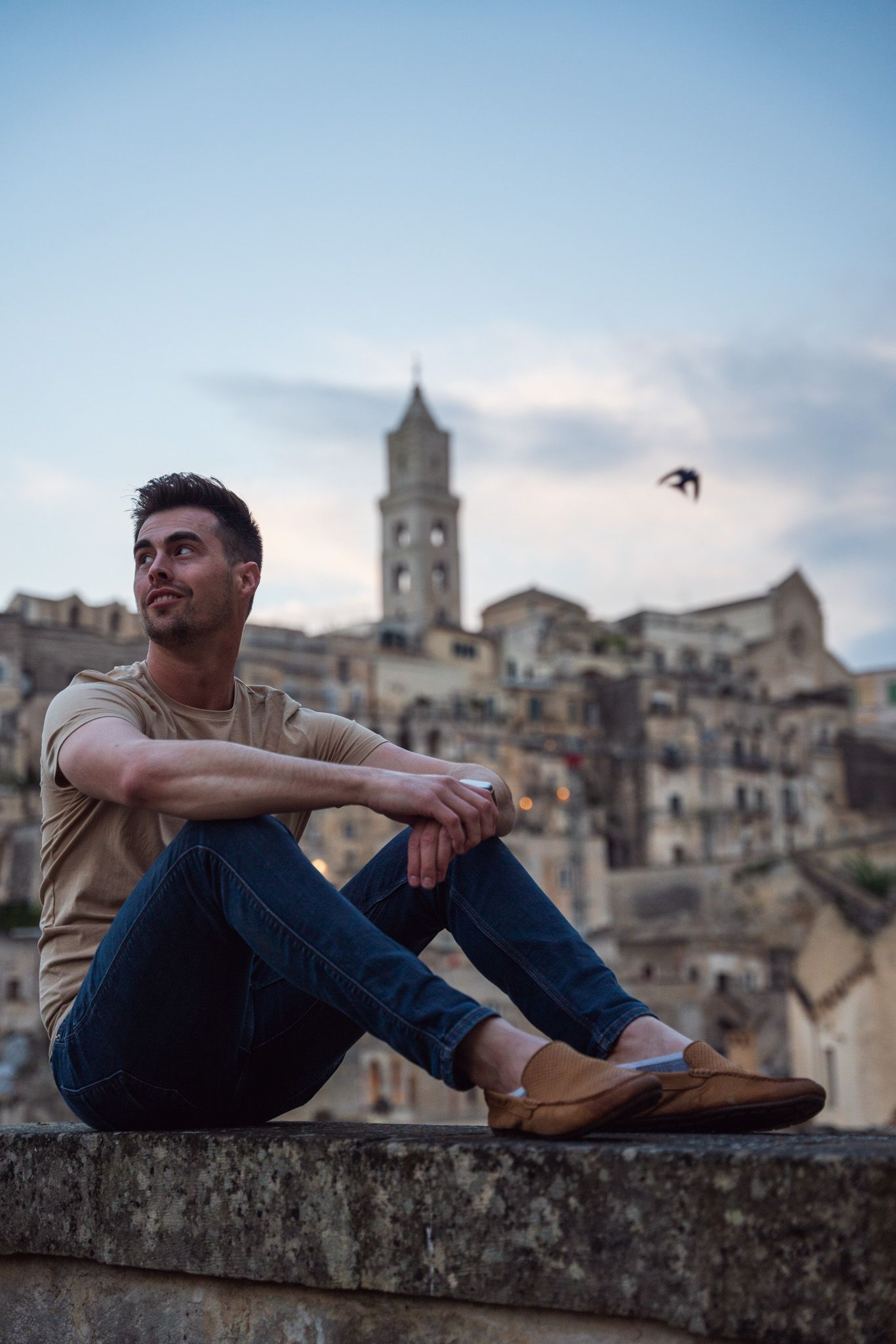Person relaxing on stone wall overlooking historic Italian town with church tower in background.