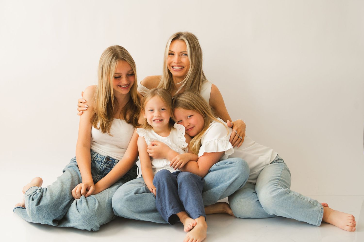 Mother and three daughters posing together in matching white tops and jeans on white background.