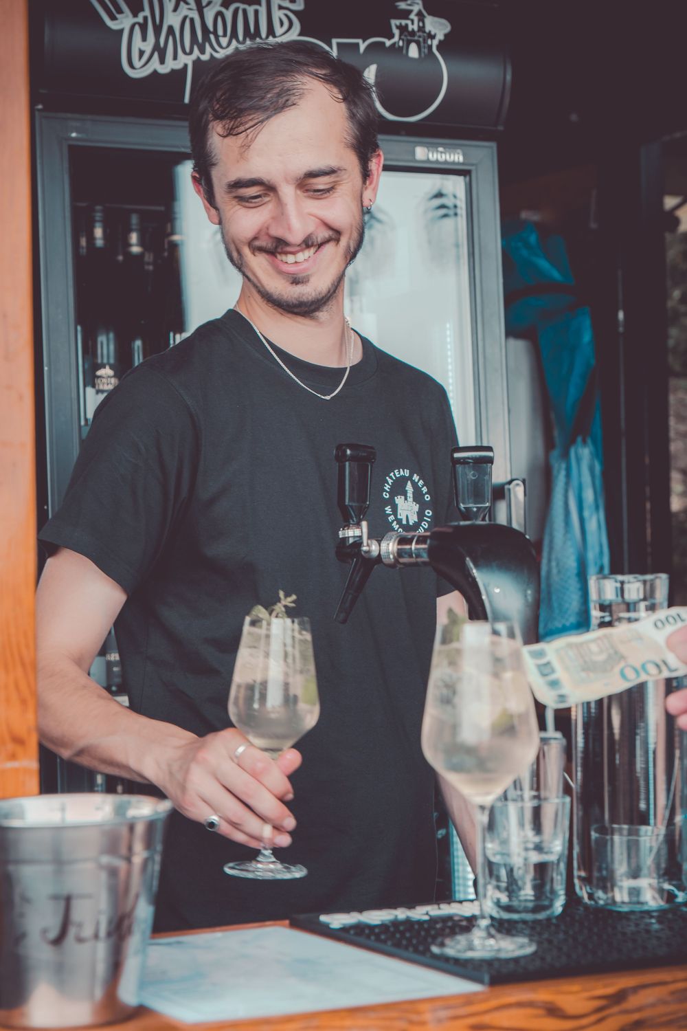 Bartender in black shirt preparing cocktails at wooden bar counter with multiple glasses.