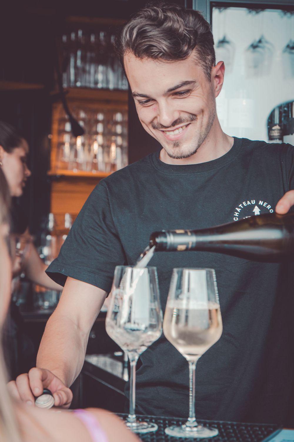 A bartender in a dark t-shirt smiles while preparing drinks at a bar counter with wine glasses.