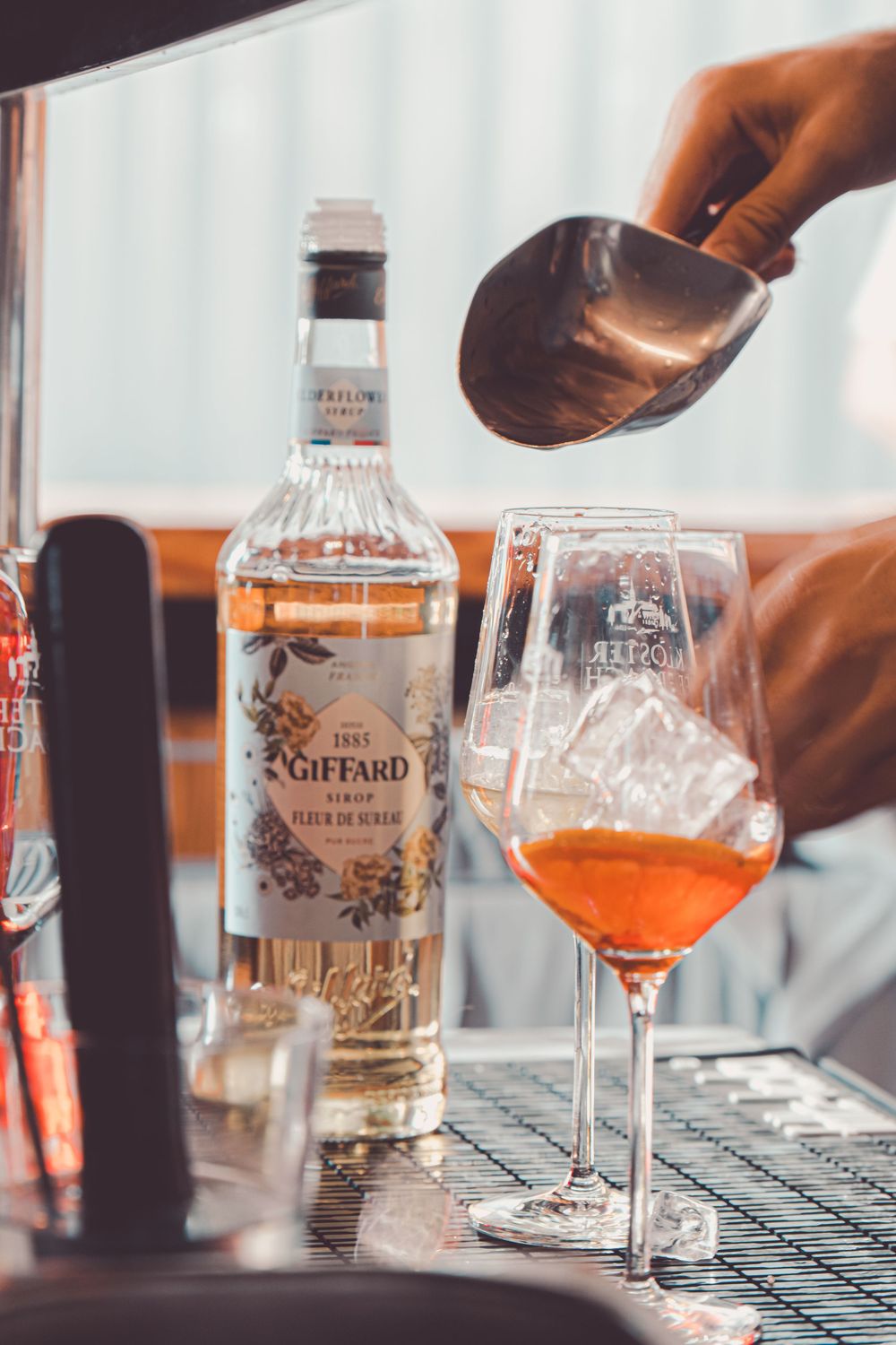 A bottle of Giffard liqueur and a wine glass being filled with orange-colored cocktail on a bar counter.
