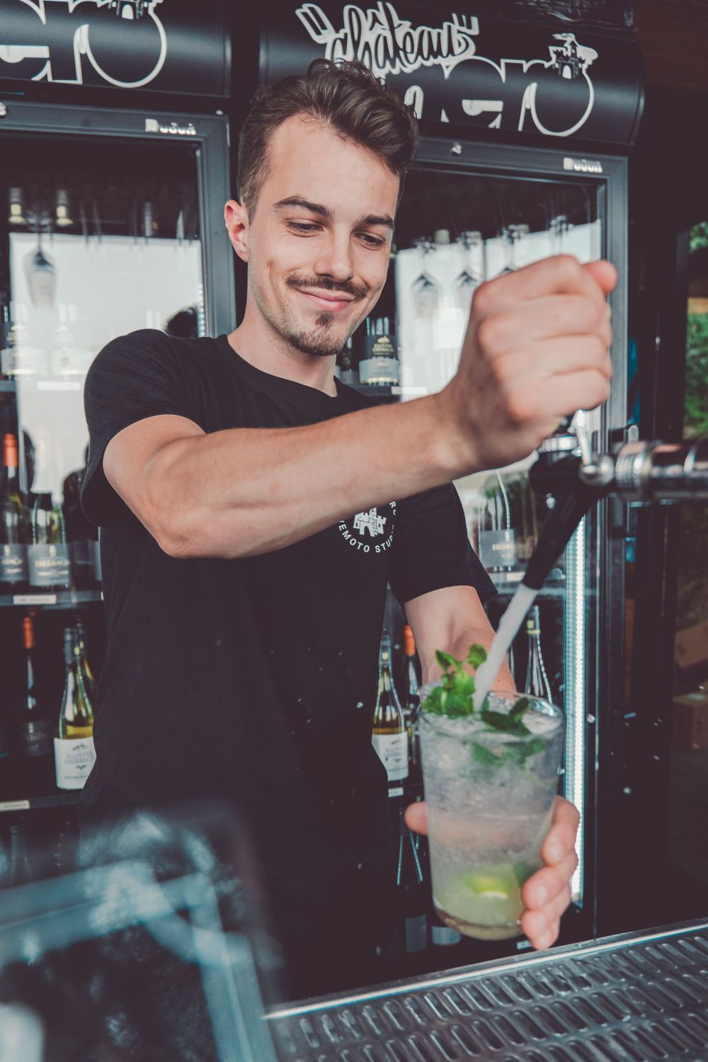 Bartender in black shirt mixes cocktails behind bar counter with focused precision and professional technique.