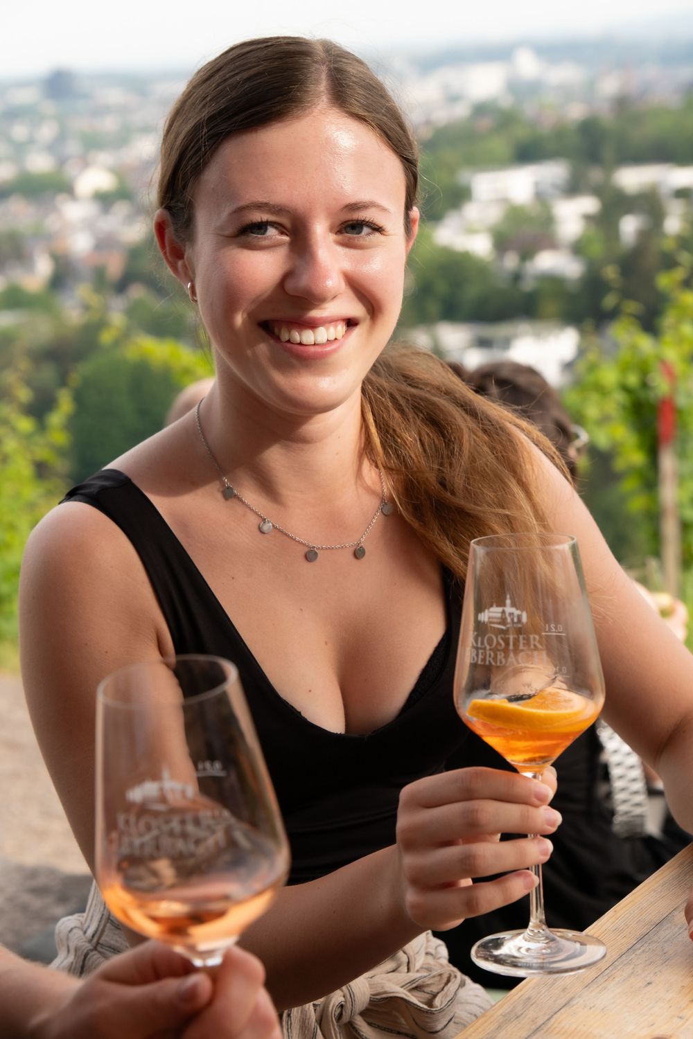 A person in a black top enjoys drinks on an outdoor patio with a scenic city view in the background.