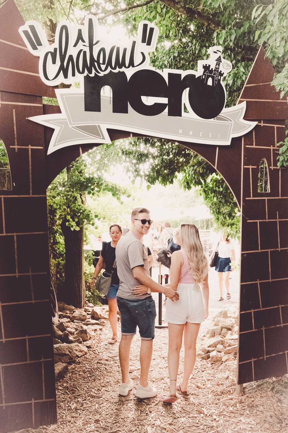 Group of people walking through a rustic outdoor pathway with brick walls and decorative signage overhead.
