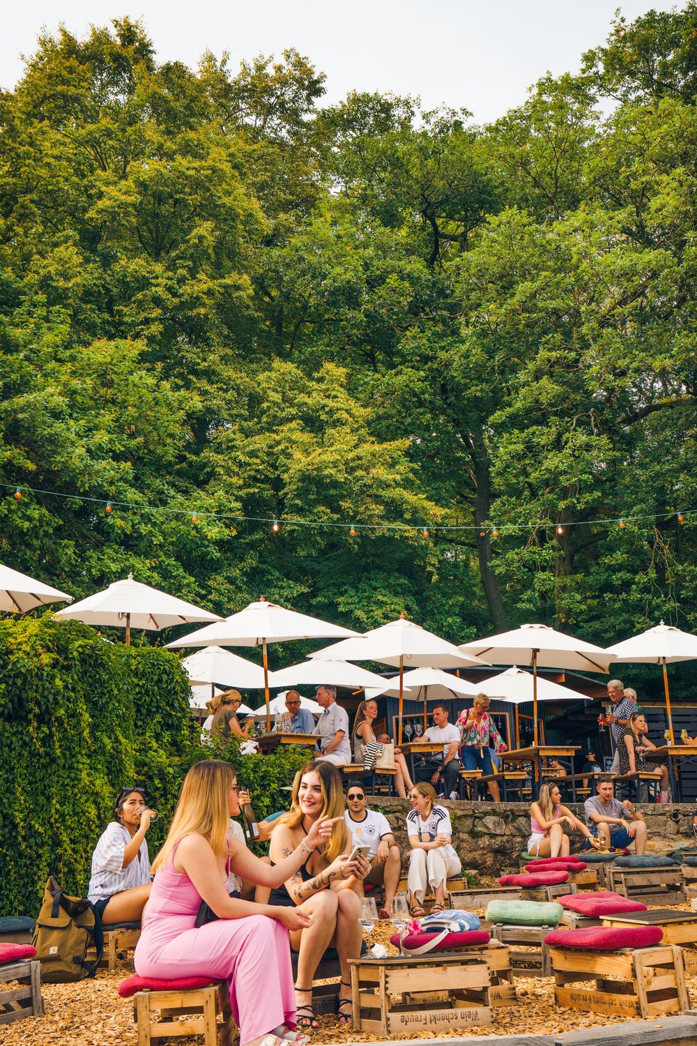 People relax on wooden pallets with cushions at an outdoor garden bar with white umbrellas and lush green trees.