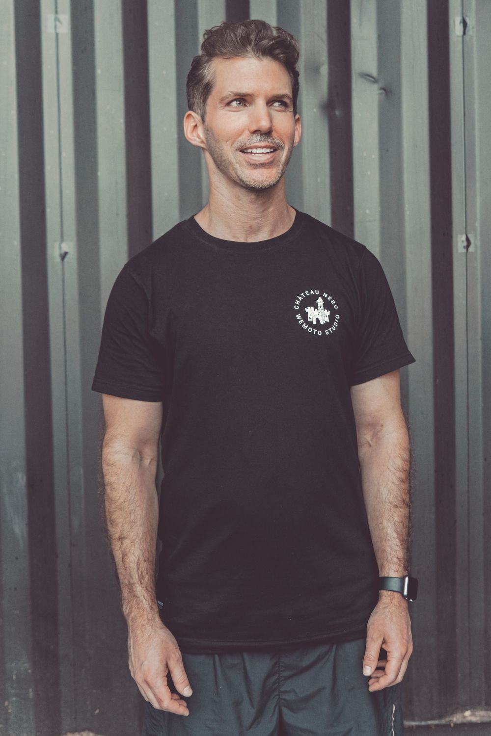 Person in dark t-shirt with small logo smiles while standing against metal wall backdrop.