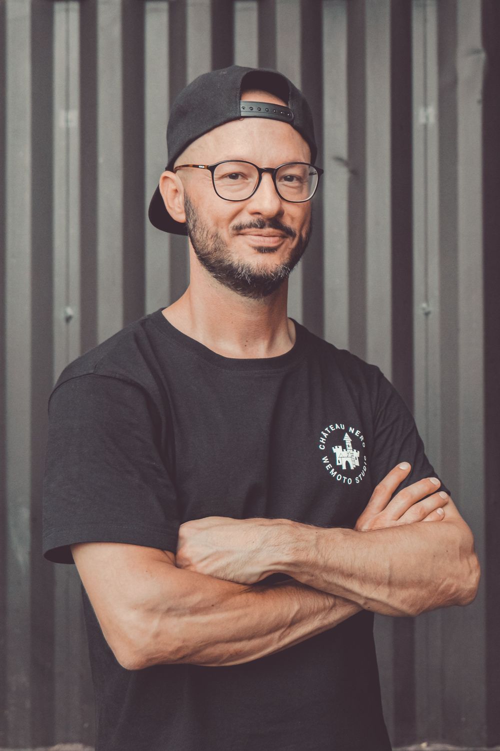 Person in black t-shirt and baseball cap with glasses stands confidently against corrugated metal wall.