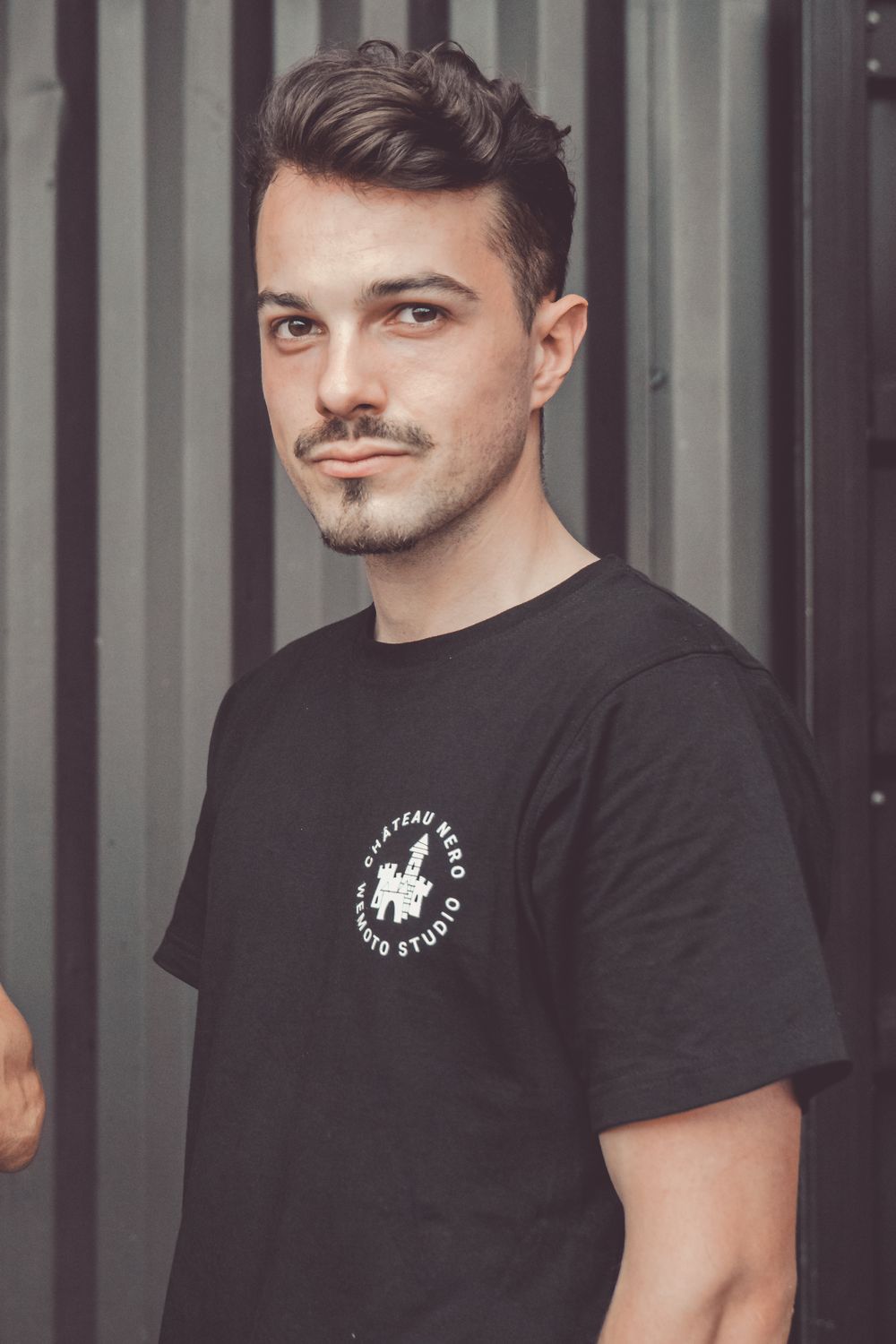 Person wearing black t-shirt with logo poses against industrial metal background.
