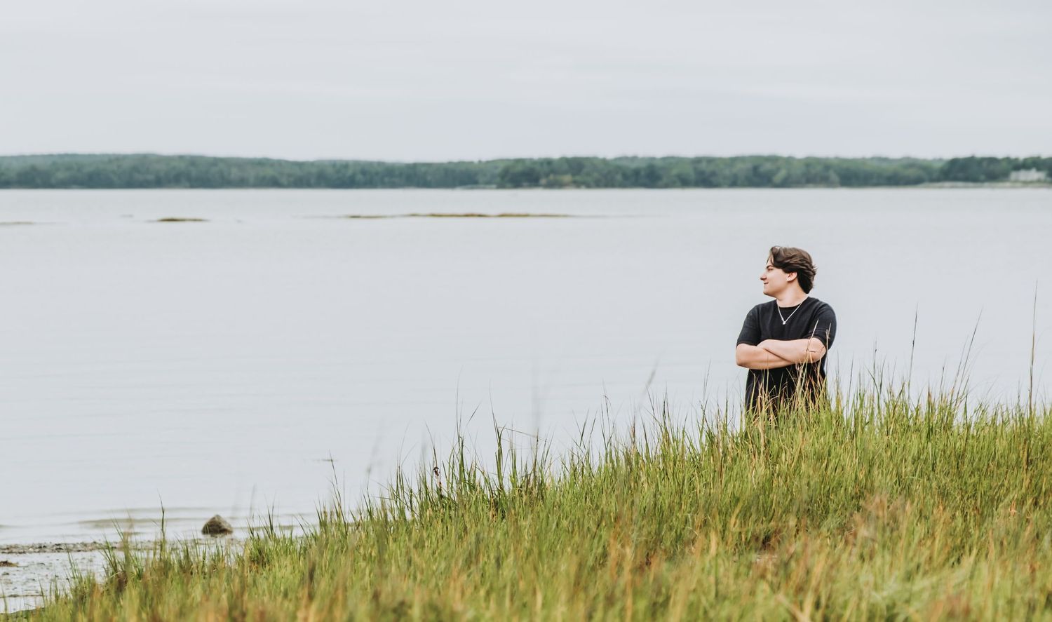 A senior in high school stands in tall grass on a hillside overlooking a serene lake or bay on an overcast day. Shot at Winslow State Park.