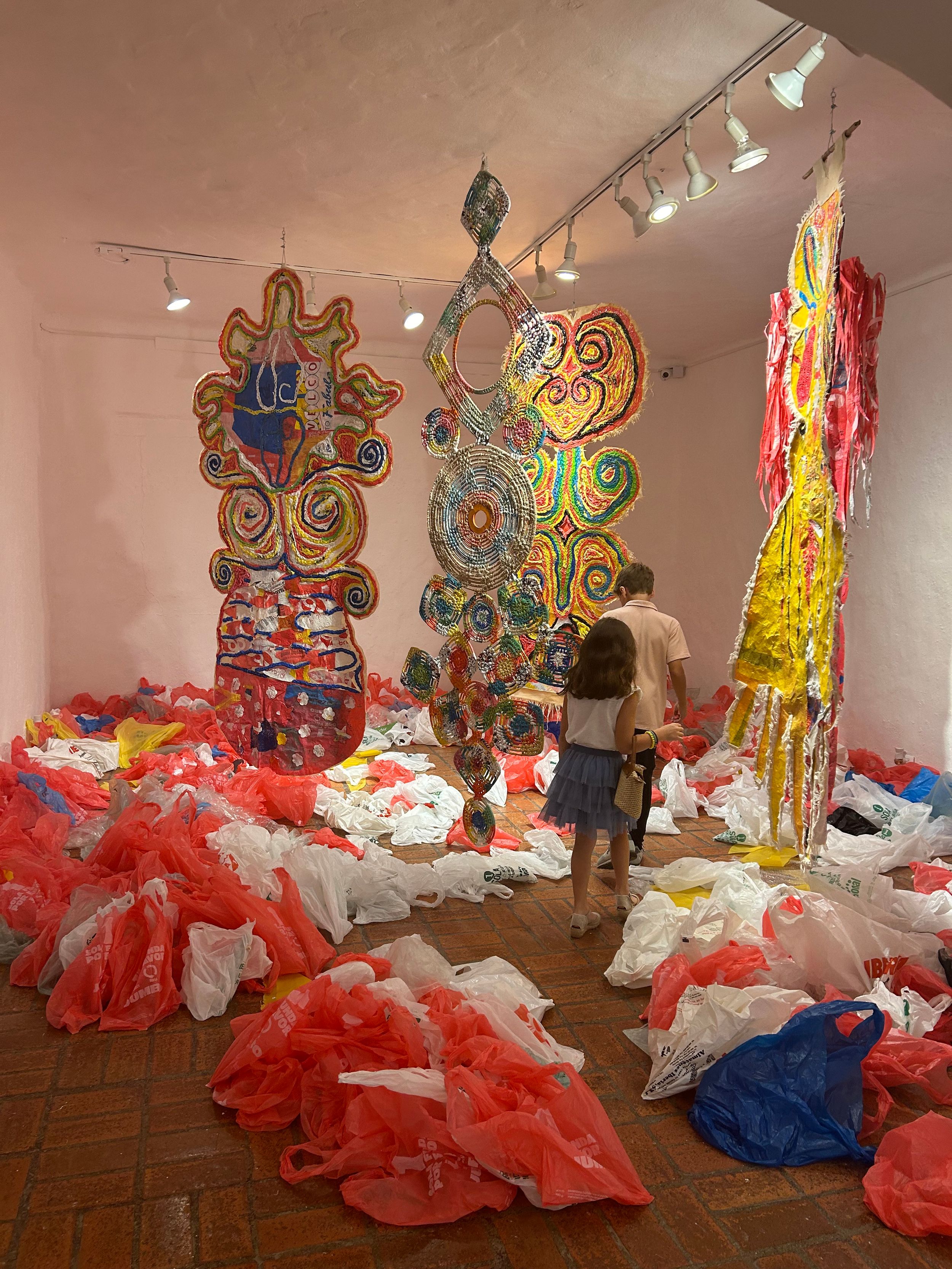 Colorful large-scale art installations hang in a gallery surrounded by scattered plastic bags, as visitors explore the vibrant exhibit.