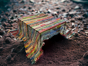 Colorful woven textile draped over a small wooden stool sitting on dark soil outdoors.