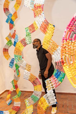 A man in black stands among towering colorful spiral sculptures made of vibrant rainbow-hued materials in a gallery.