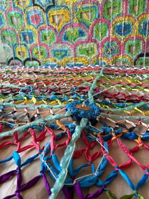 Colorful handmade net craft with knotted multicolored ribbons, displayed against a vibrant embroidered textile background.