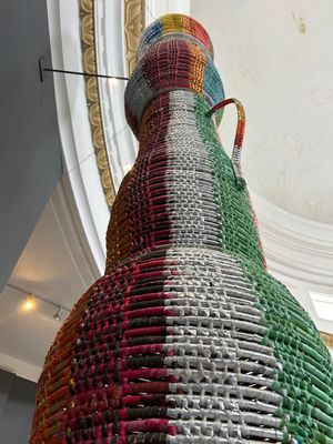 Towering colorful woven sculpture made from recycled materials displayed in a museum atrium, viewed from below.