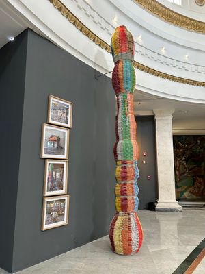 Colorful woven textile tower sculpture displayed in a rotunda gallery alongside framed artwork prints on a gray wall.