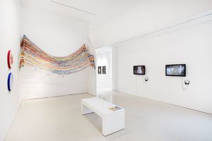 Modern art gallery with colorful textile wall installation, white bench, video screens, and minimalist white walls.