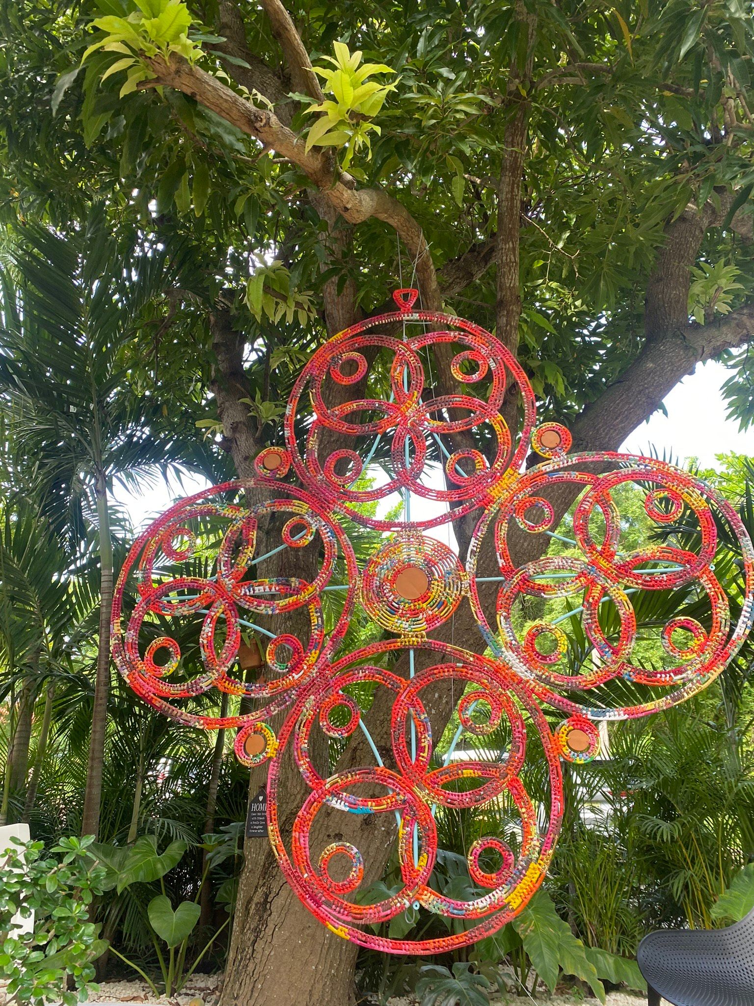 Red ornate metal cross wind spinner hanging from a tropical tree in a lush garden setting.