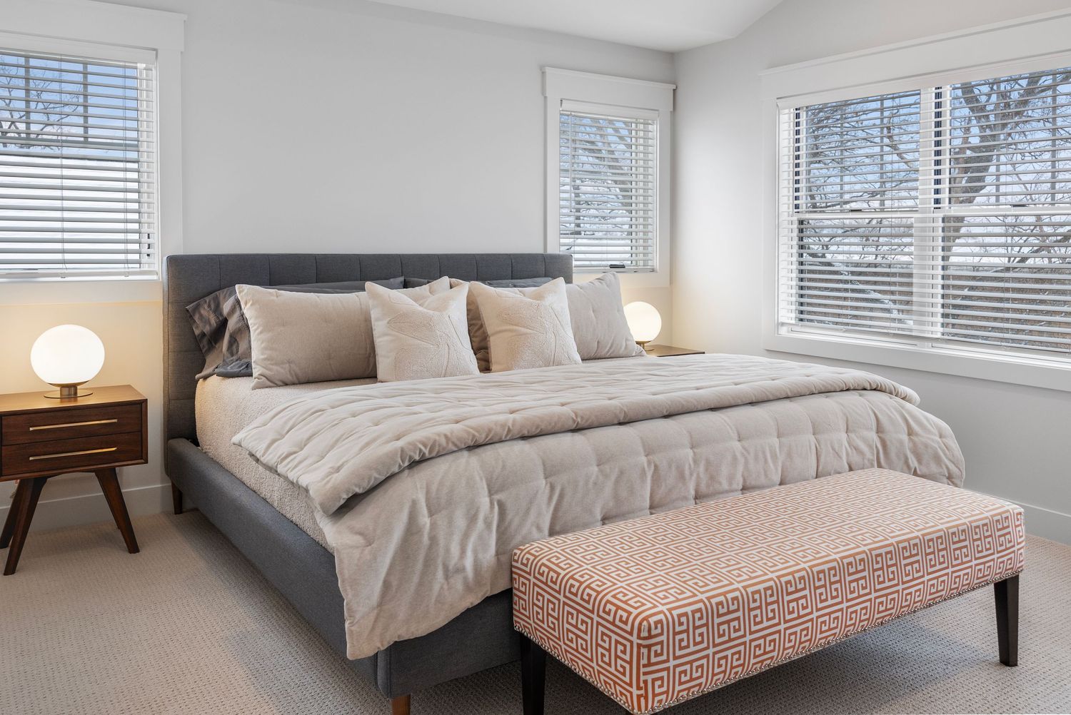 Modern bedroom with grey bed, pink bench, and natural light from three windows with blinds.