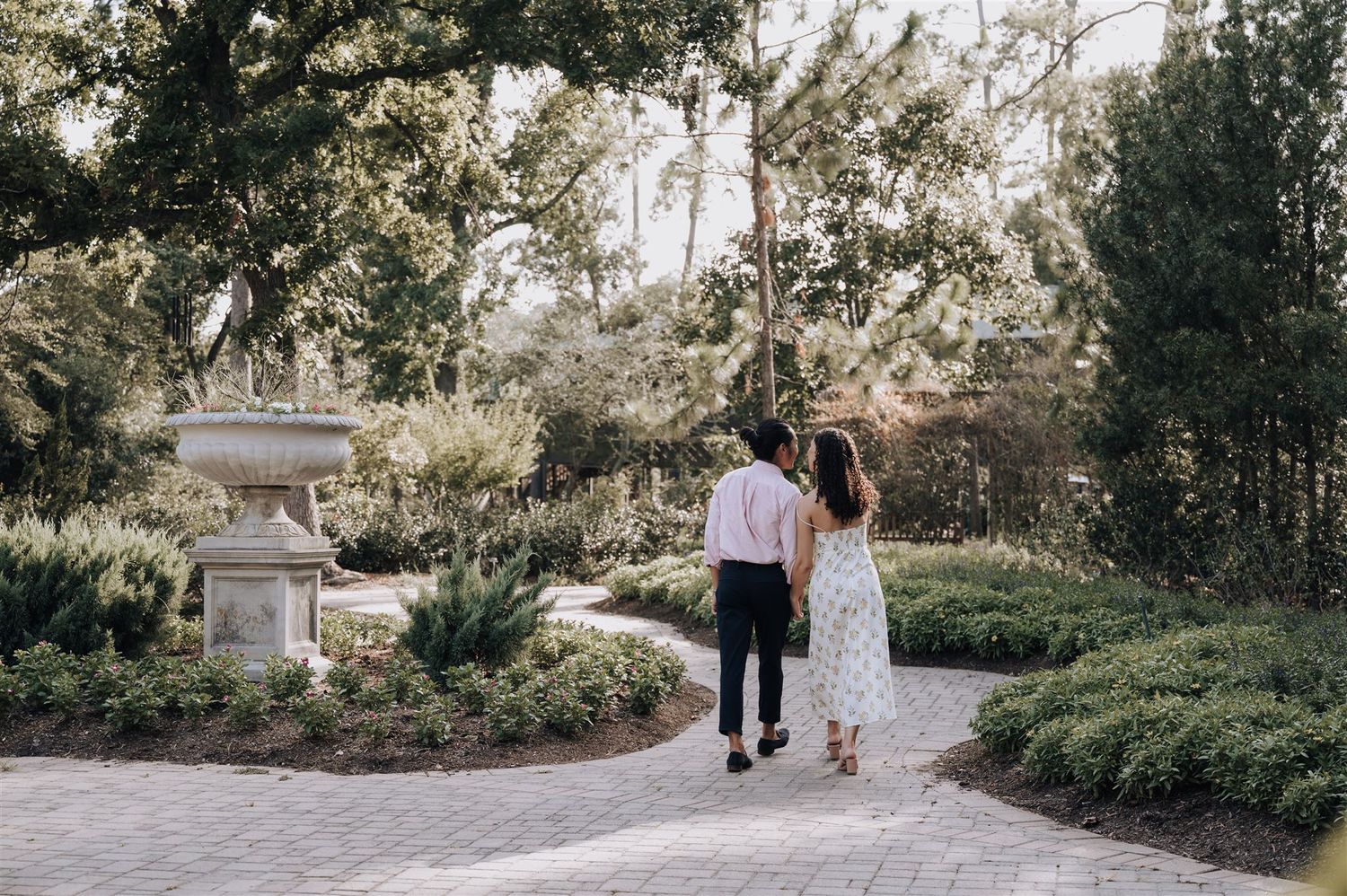 A couple walks hand in hand along a garden path surrounded by lush greenery and a stone fountain.