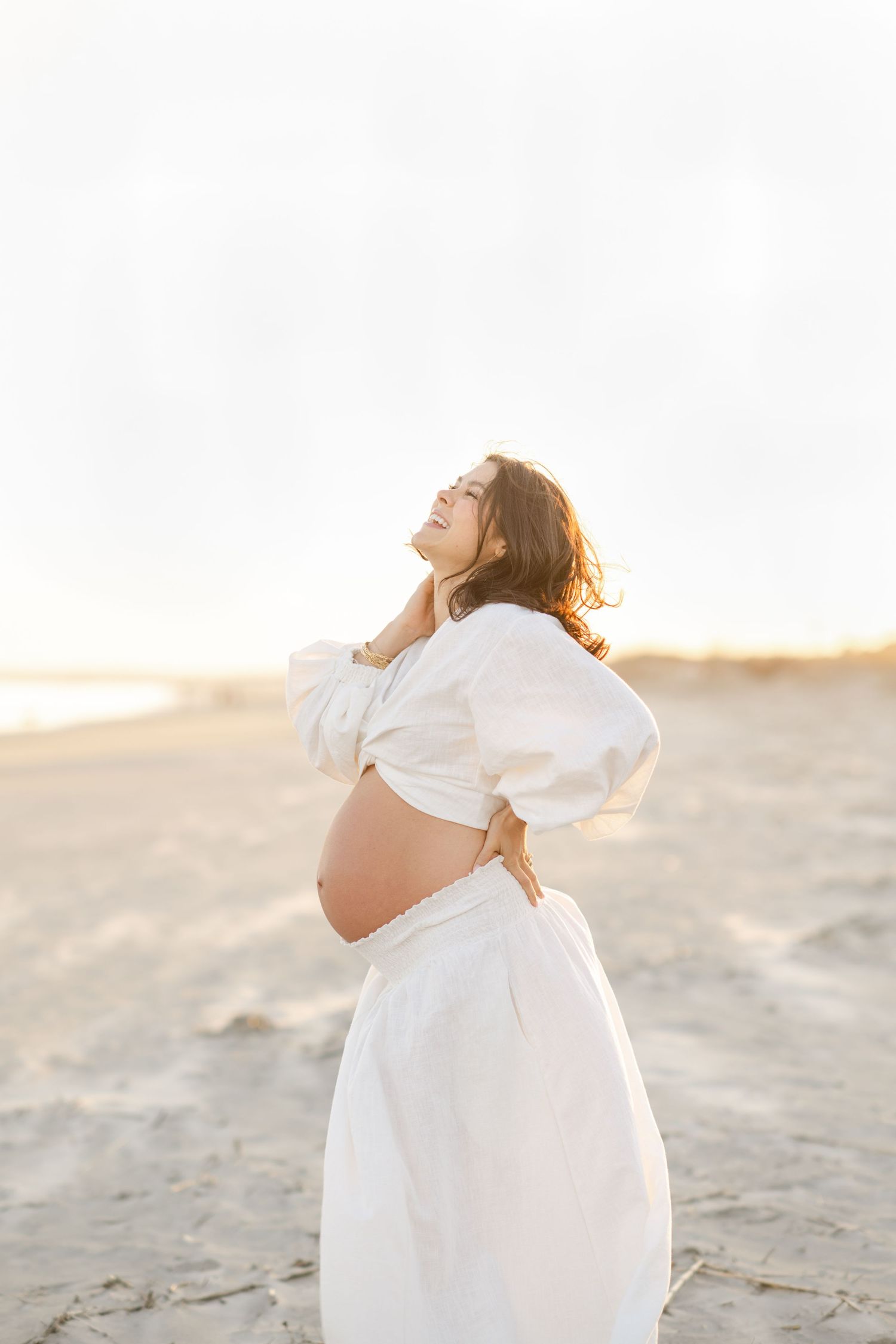 A joyful pregnant woman in white crop top and skirt poses on a sandy beach at golden hour.