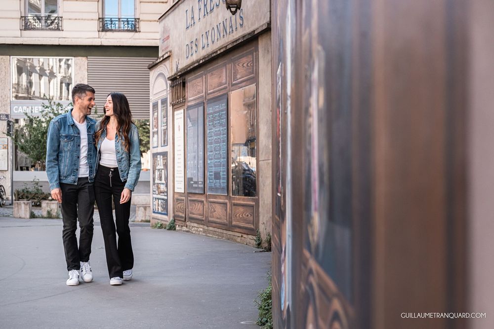 Couple holding hands and smiling at each other on a Parisian street in front of a vintage French storefront.
