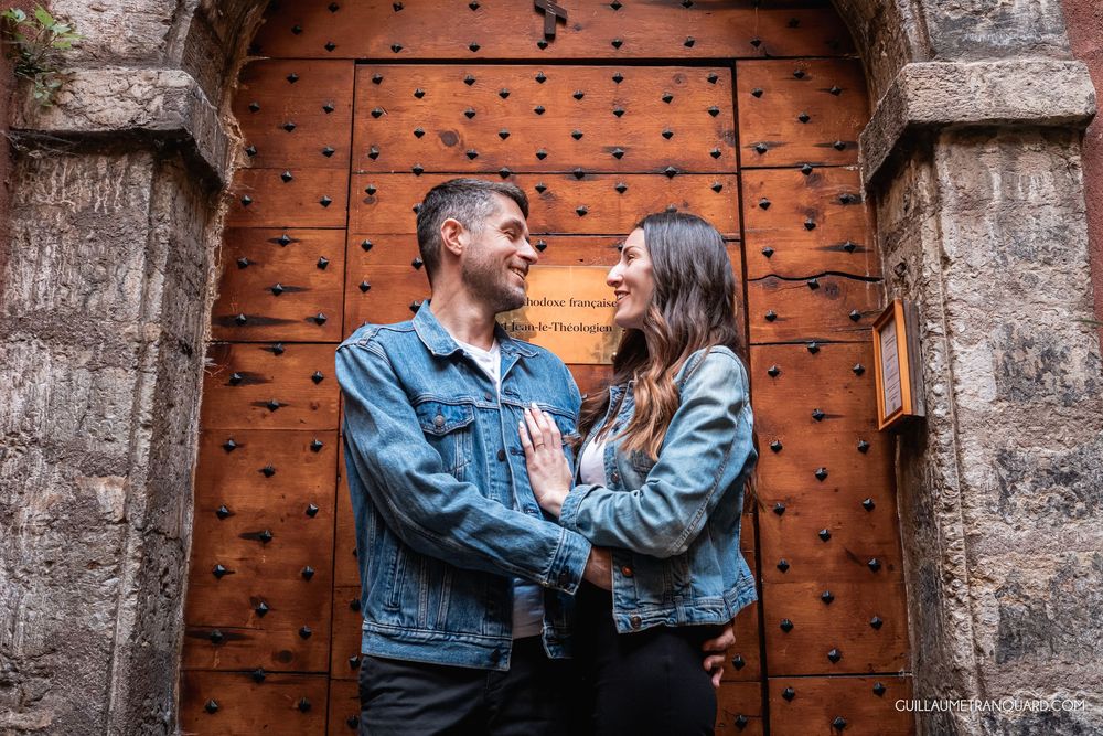 Couple in matching denim jackets smiling at each other in front of a rustic wooden door with metal studs.