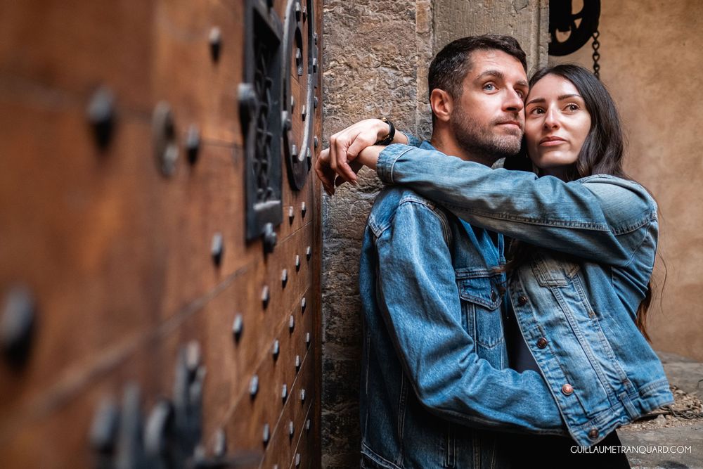 Couple in denim jackets leaning against an ancient stone wall with ornate wooden door.
