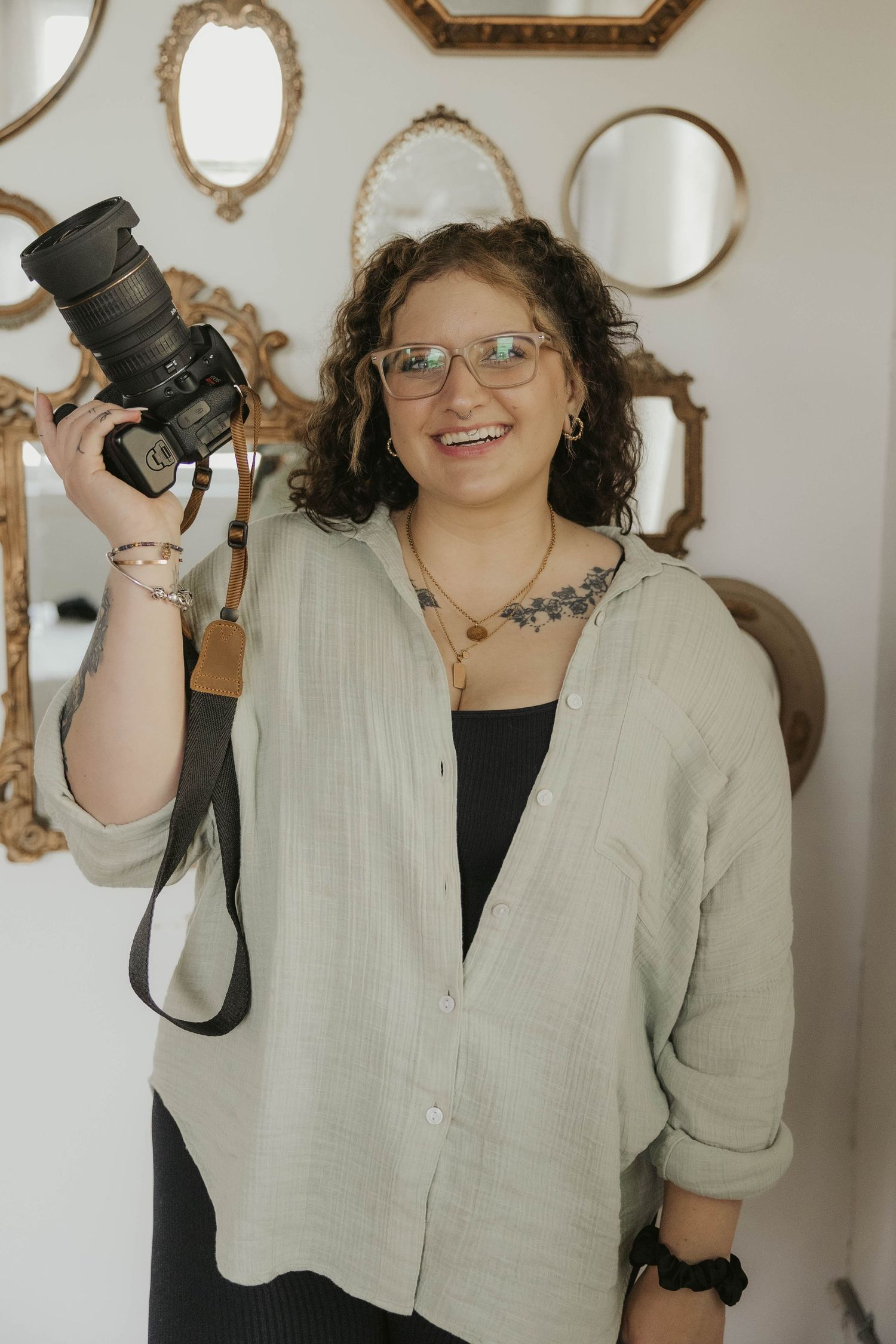 Professional photographer poses with camera equipment in front of ornate wall mirrors.