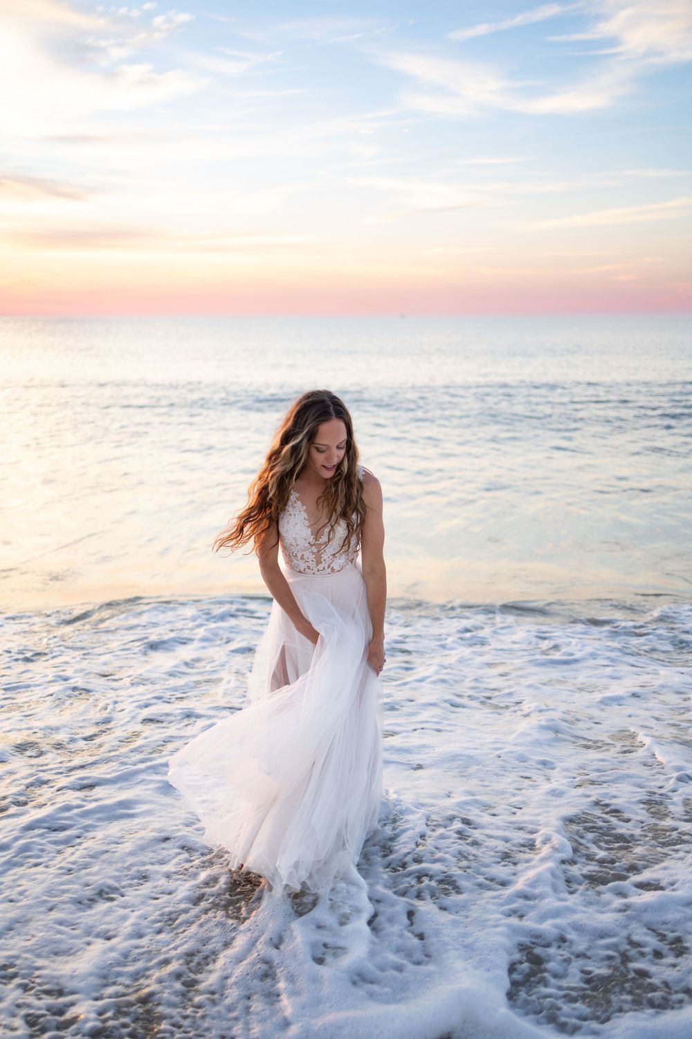A bride in a flowing white lace gown stands in ocean waves at sunset, her hair blowing in the breeze.