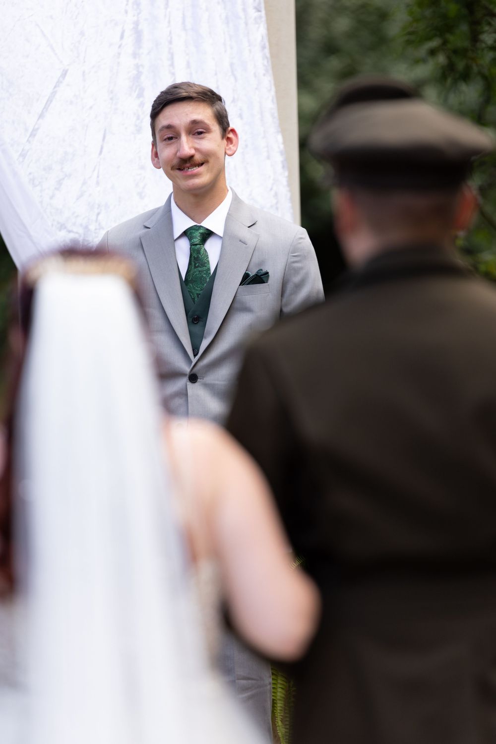 A groom in gray suit with green tie stands at wedding ceremony altar during outdoor wedding.