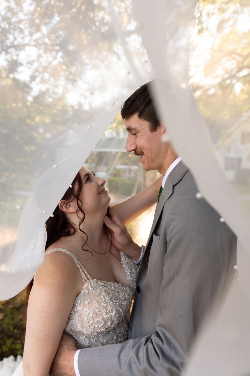 A couple in wedding attire share an intimate moment together against a sunlit outdoor backdrop with dreamy bokeh effects.