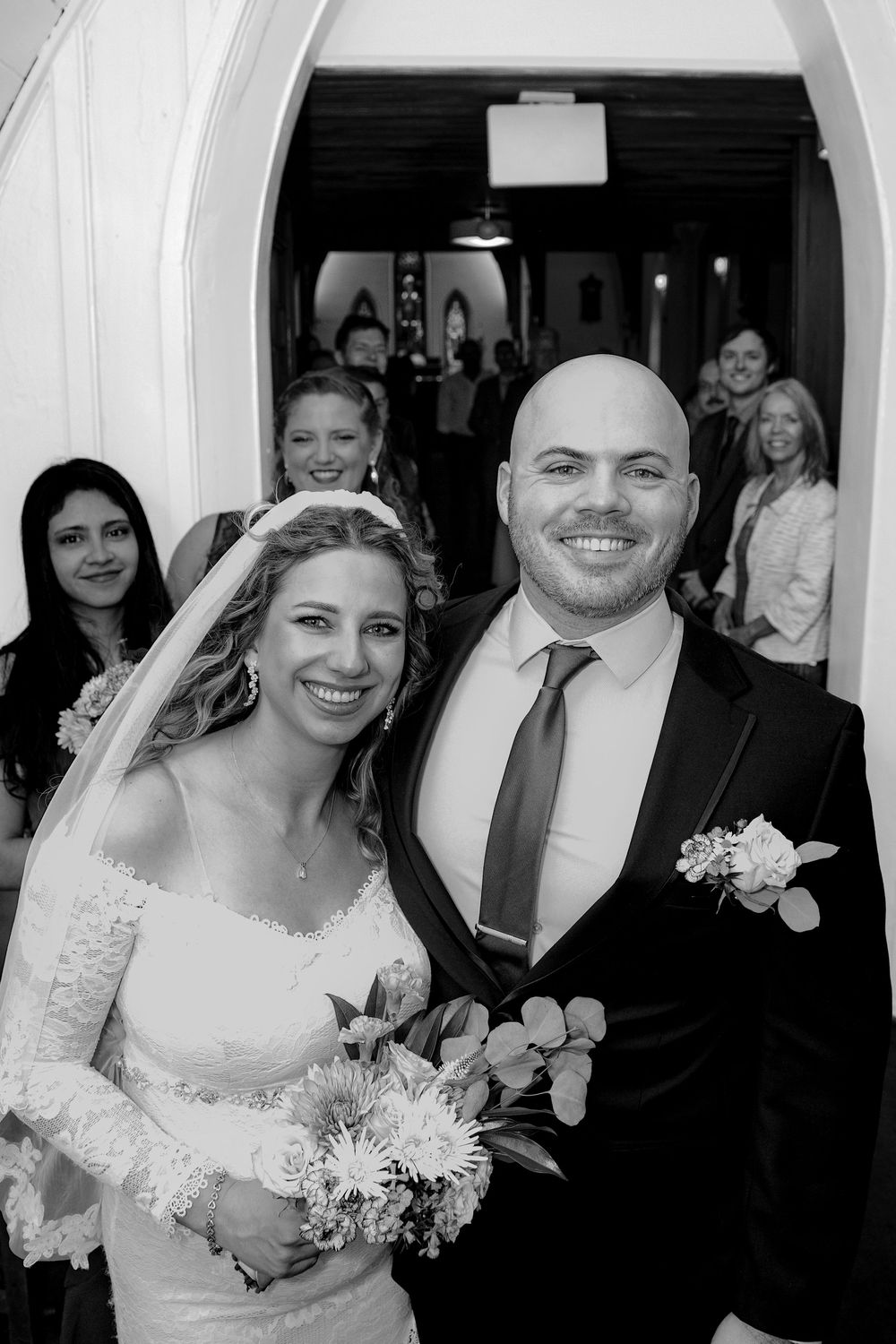 A joyful black and white wedding photo showing a couple in traditional wedding attire with guests in the background.