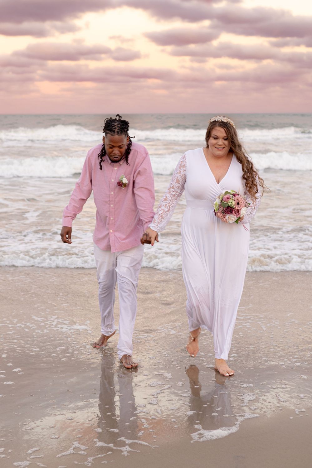 Couple walking together on beach at sunset, bride holding bouquet.