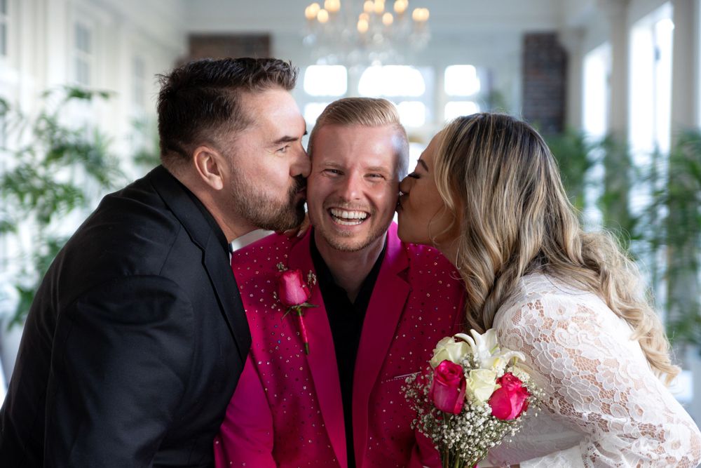 Three people share a joyful moment at a wedding with red and white floral bouquet details.