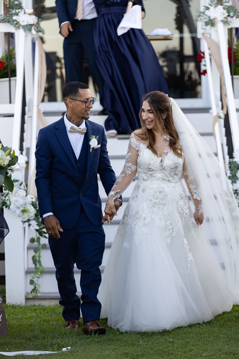 A bride in a white lace gown and groom in navy suit walk hand in hand on grass by white floral decorated stairs.