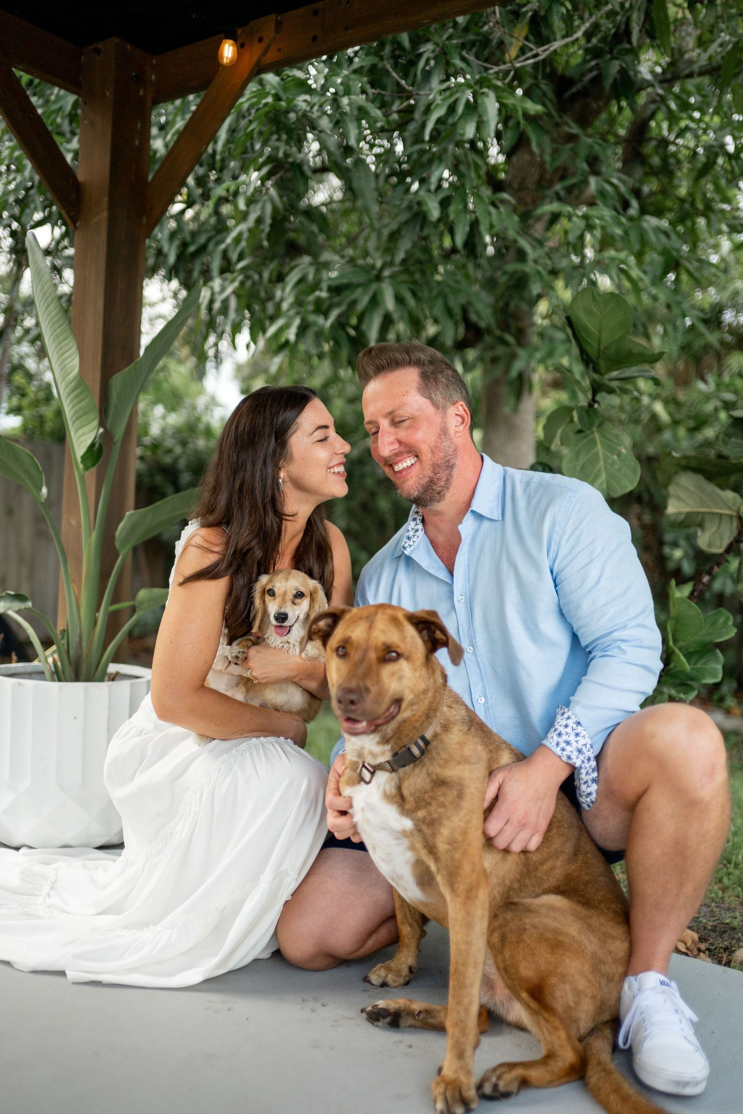 Couple sitting on porch with their brown dog under lush green foliage.