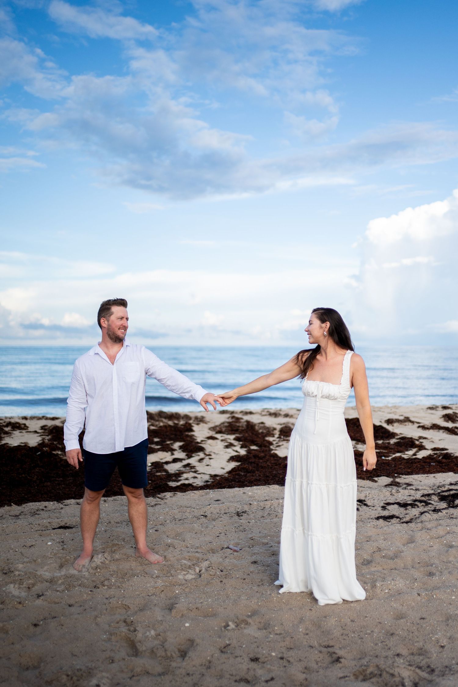 Couple in white clothing holding hands on a rocky beach under blue skies.