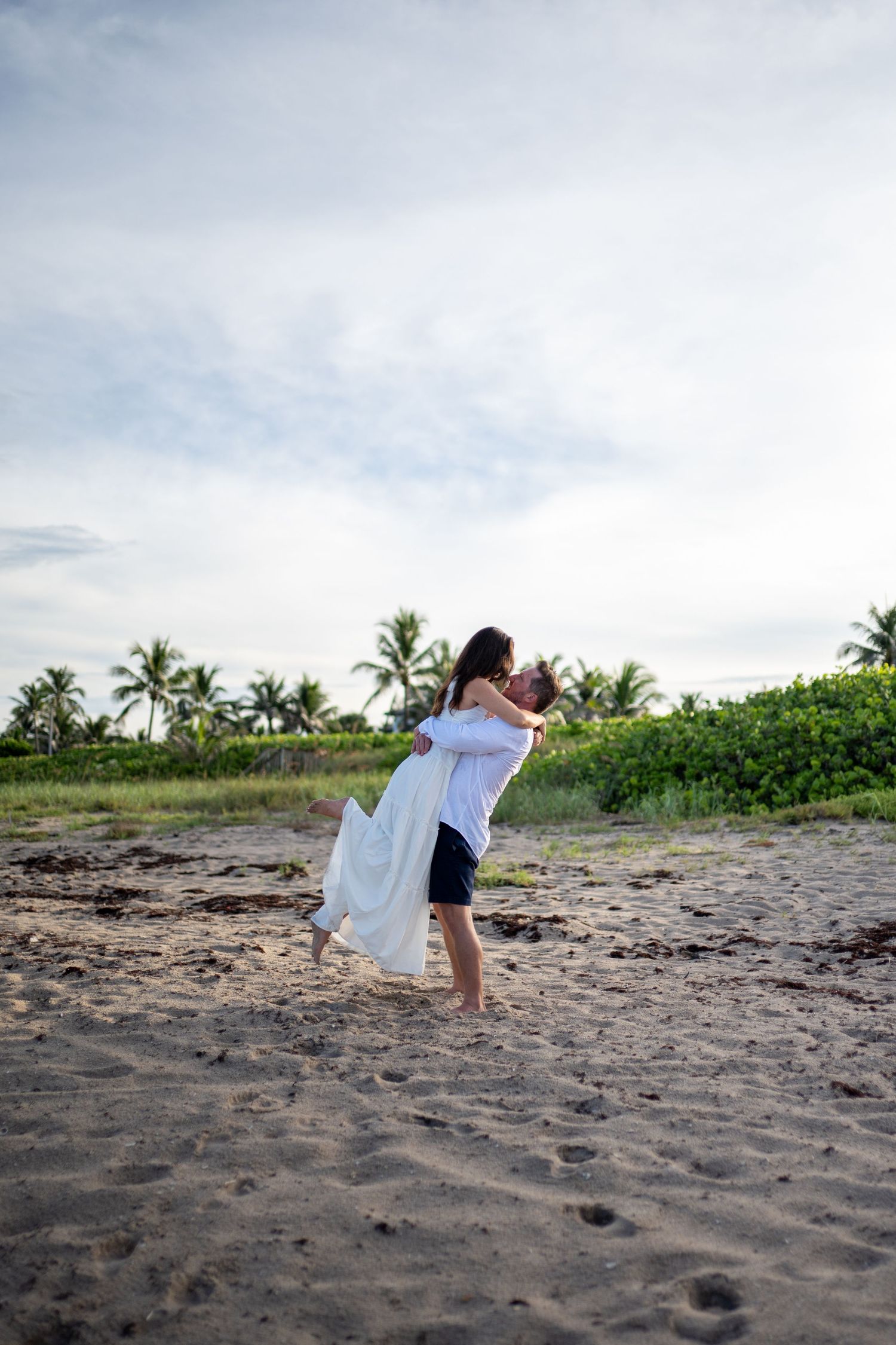 Couple in white outfits sharing intimate moments on a tropical beach with palm trees in background.