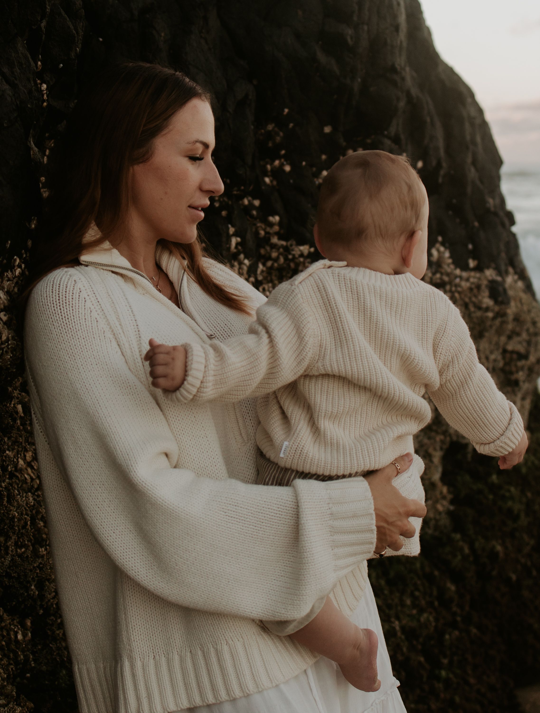 Mother and baby in matching cream sweaters share a tender moment against a rocky coastal backdrop at sunset.